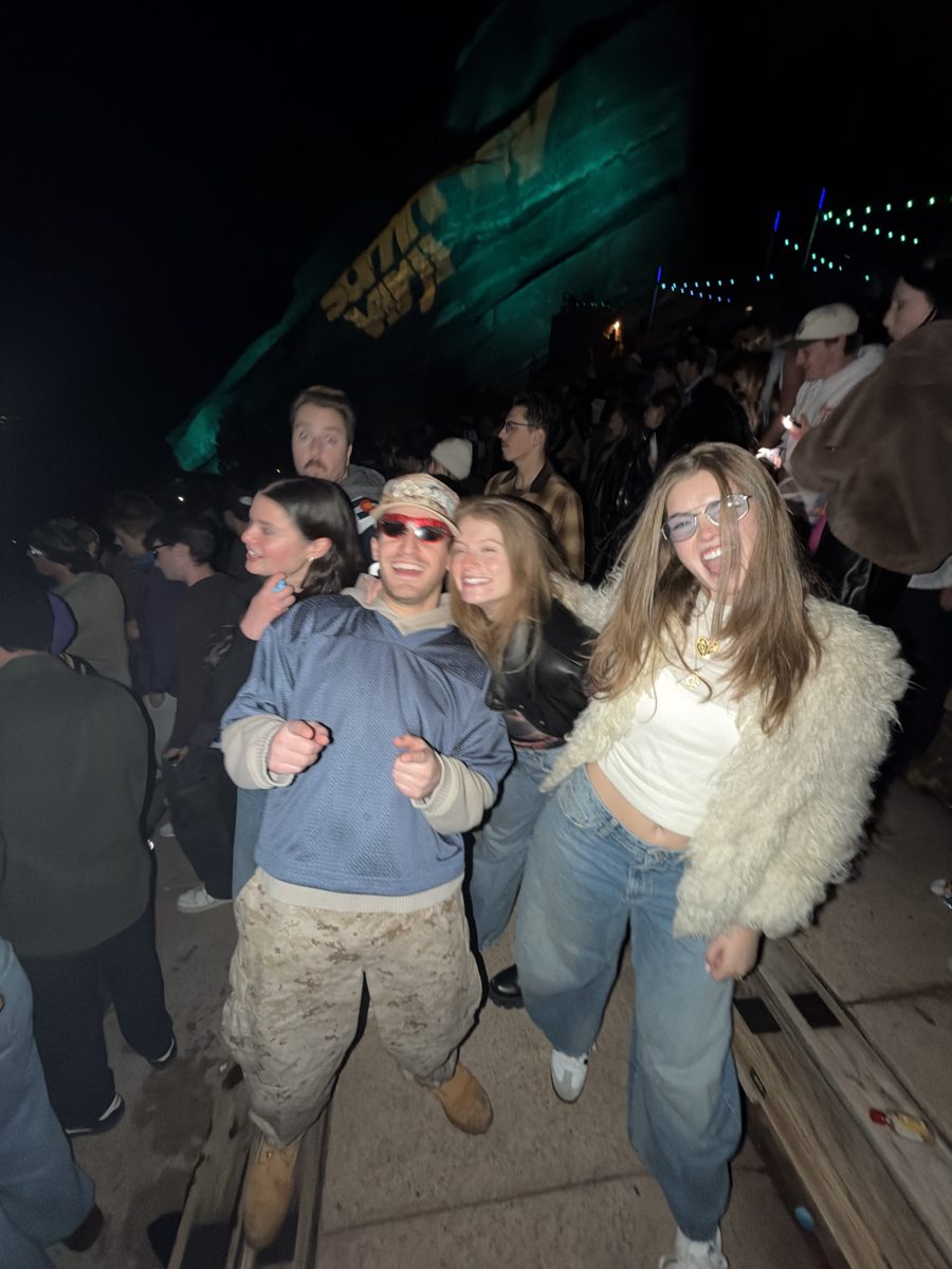 The crew dancing on the Red Rocks rows at night: Simi on the left with dark hair, Zack in the center in a blue jersey and camo pants, Dalia behind him in a leather jacket, and Molly on the right in a fur coat with her mouth open. The rocks glow green behind them.